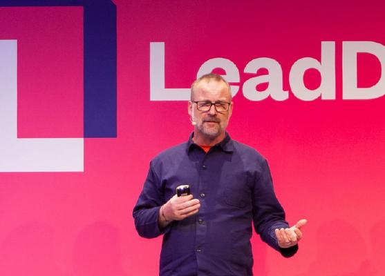Joel wearing a blue jacket, againt a pink background speaking on stage